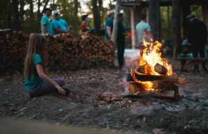 A young girl sits by a campfire in a forest setting with a group of people in the background.